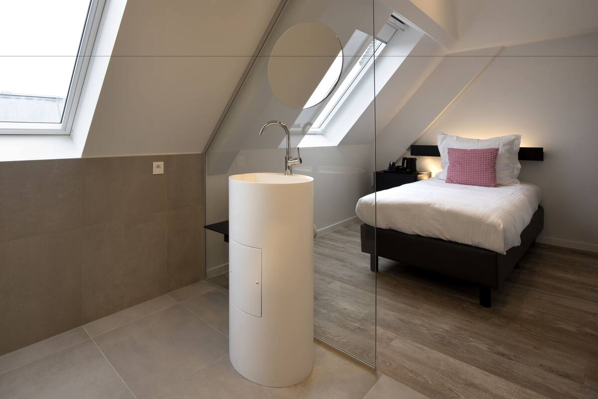 A modern bedroom with a skylight, a round white sink, a bed with white bedding, and a nightstand with a lamp.