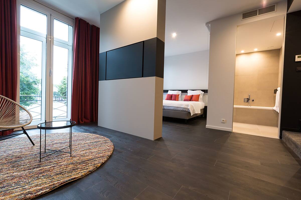 A modern hotel room with a large bed, a separate bathroom, a seating area with a chair and a small table, and a view through a glass door.