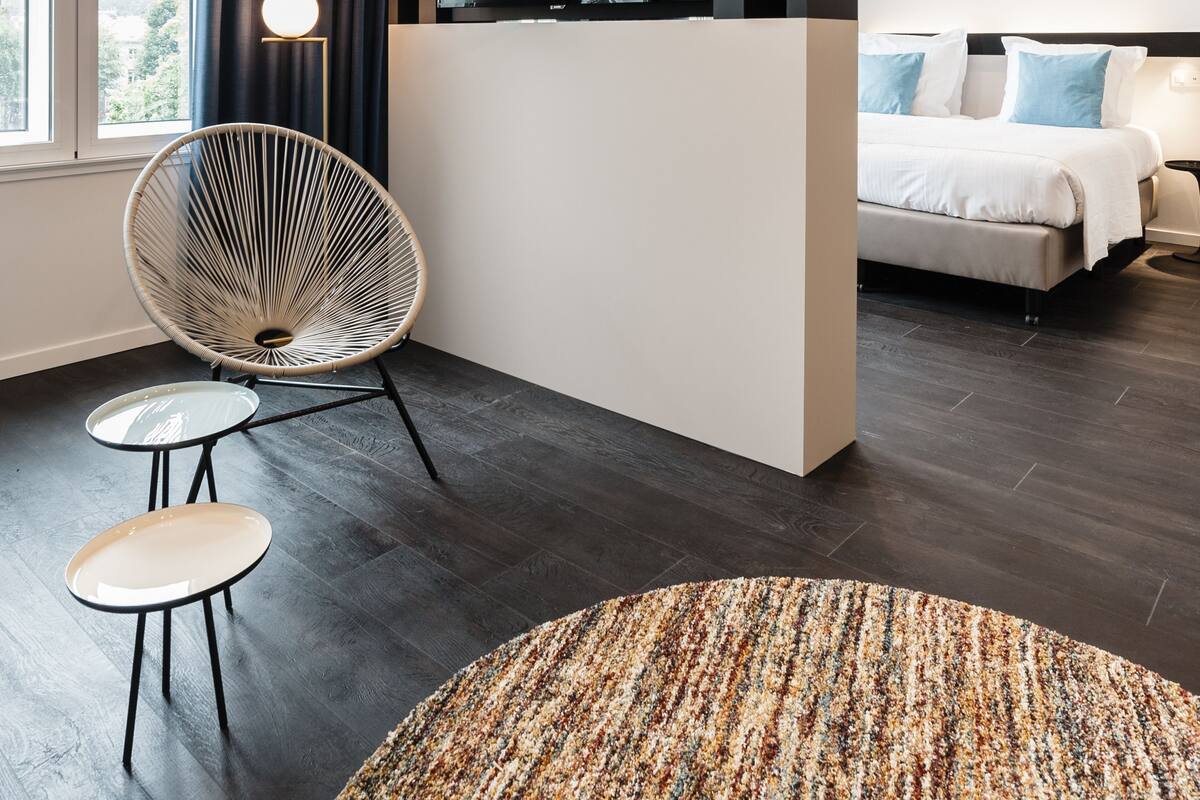 A modern hotel room with a large bed, a flat-screen TV mounted on the wall, a round chair, a small table, and a patterned rug.