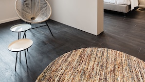 A modern hotel room with a large bed, a flat-screen TV mounted on the wall, a round chair, a small table, and a patterned rug. - Hotel Portinari (Bruges)