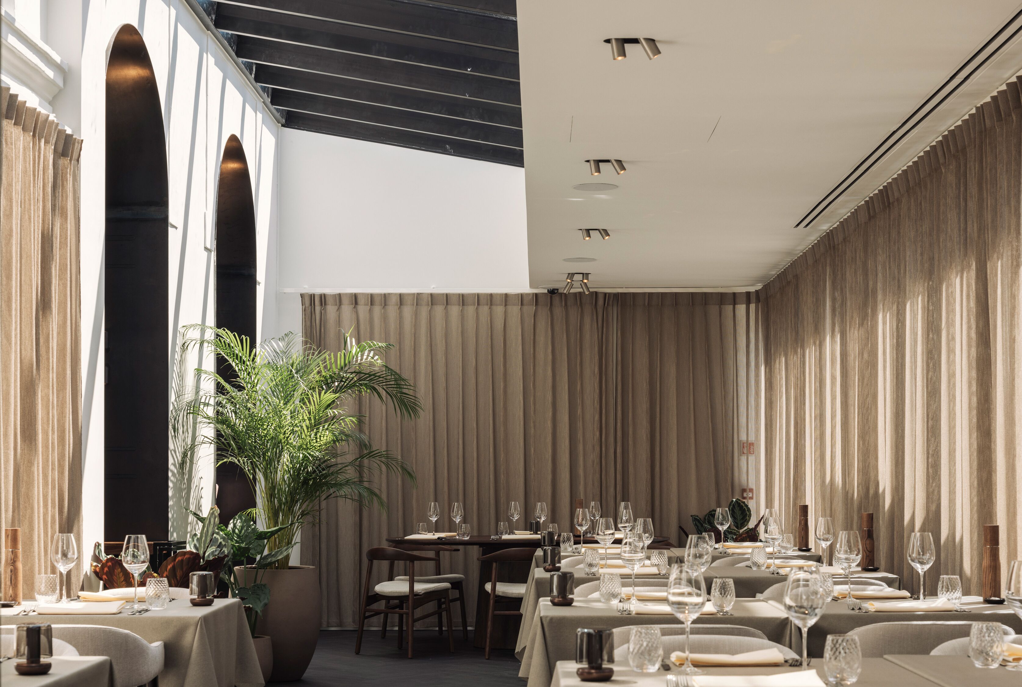 Een restaurant met meerdere tafels gedekt, grote potplanten en een modern interieur.