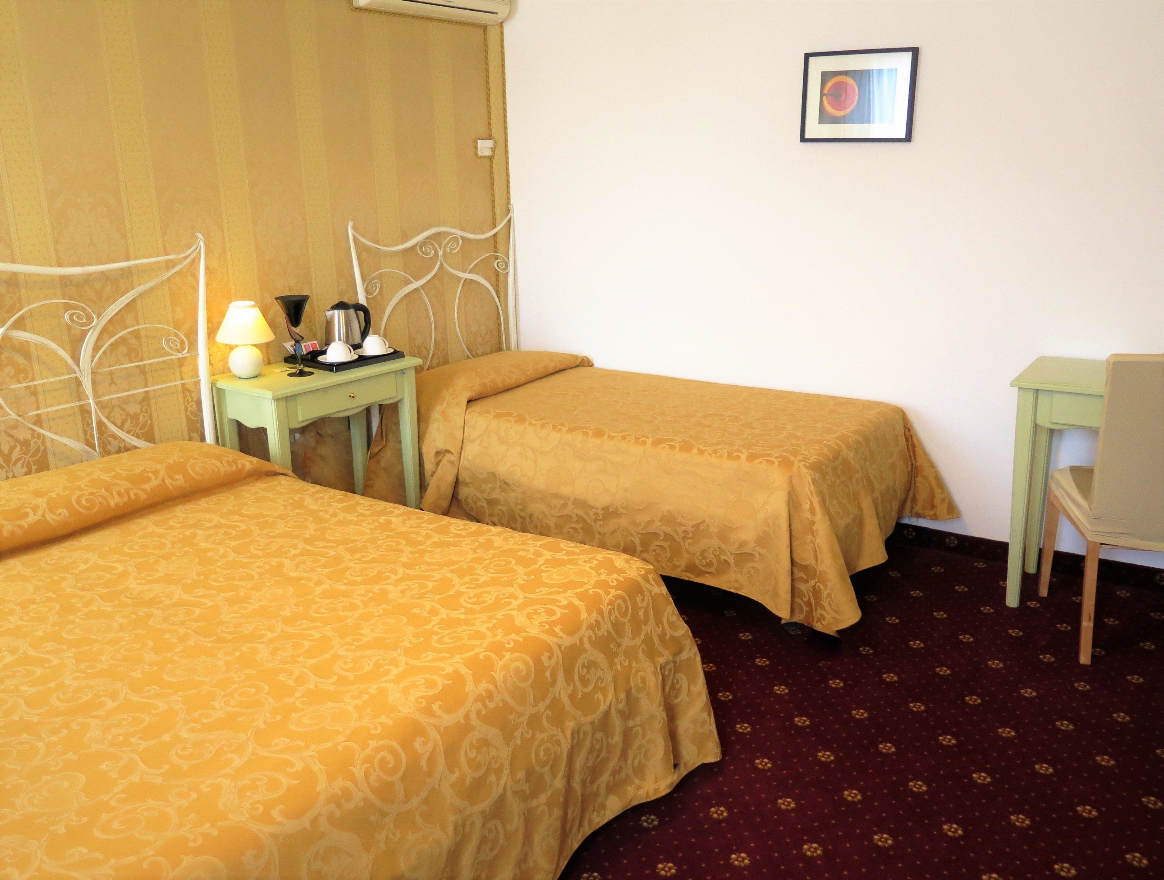 A hotel room with two single beds, a small table with a lamp, a chair, and a framed picture on the wall.