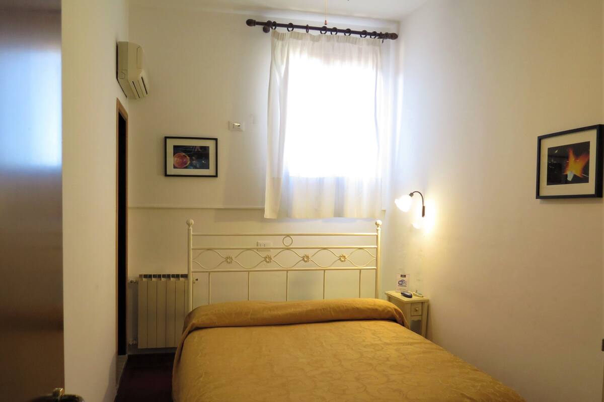A hotel room with a bed, a bedside table, a nightstand, a wall-mounted light, a window with curtains, and a radiator.