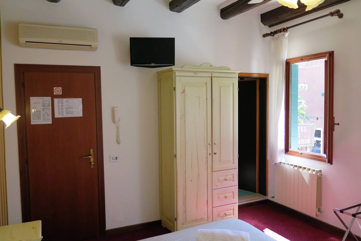 A room with a bed, a wardrobe, a wall-mounted TV, a door with a phone, and a window with a view of the outside.