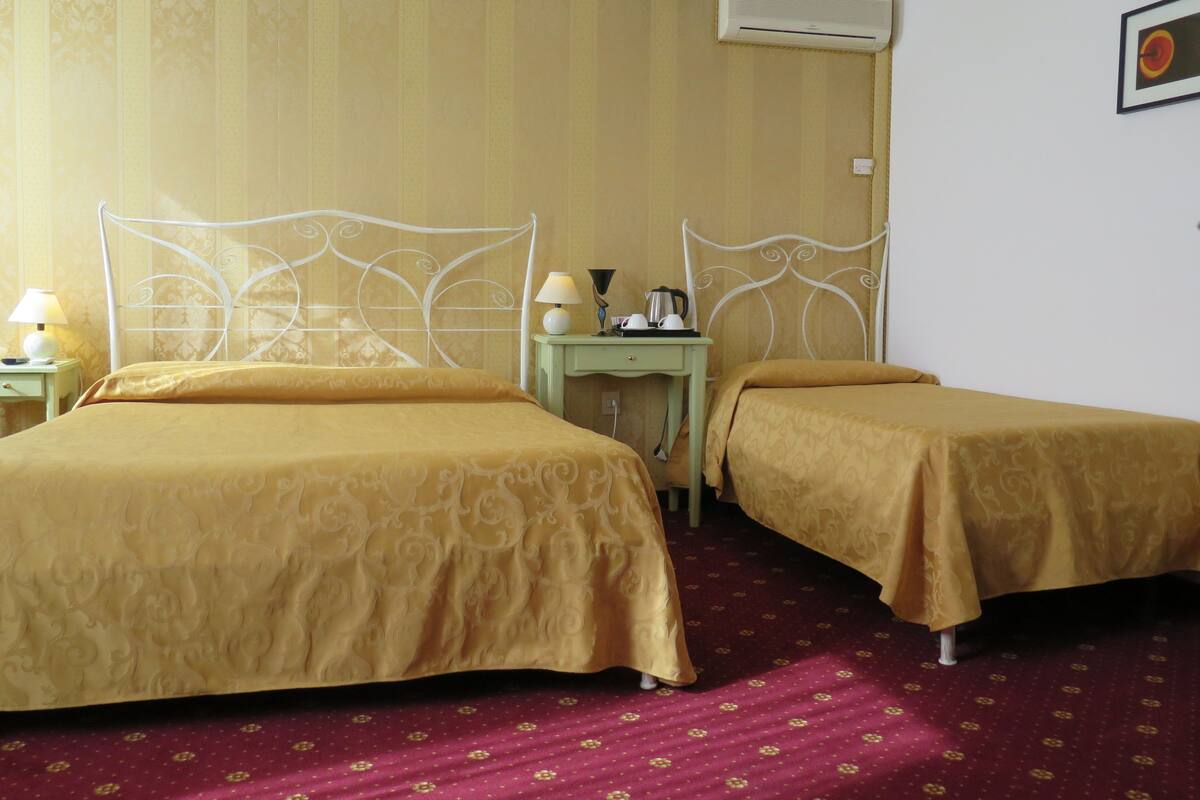 A hotel room with two beds, a nightstand, a table with a lamp, and an air conditioning unit.