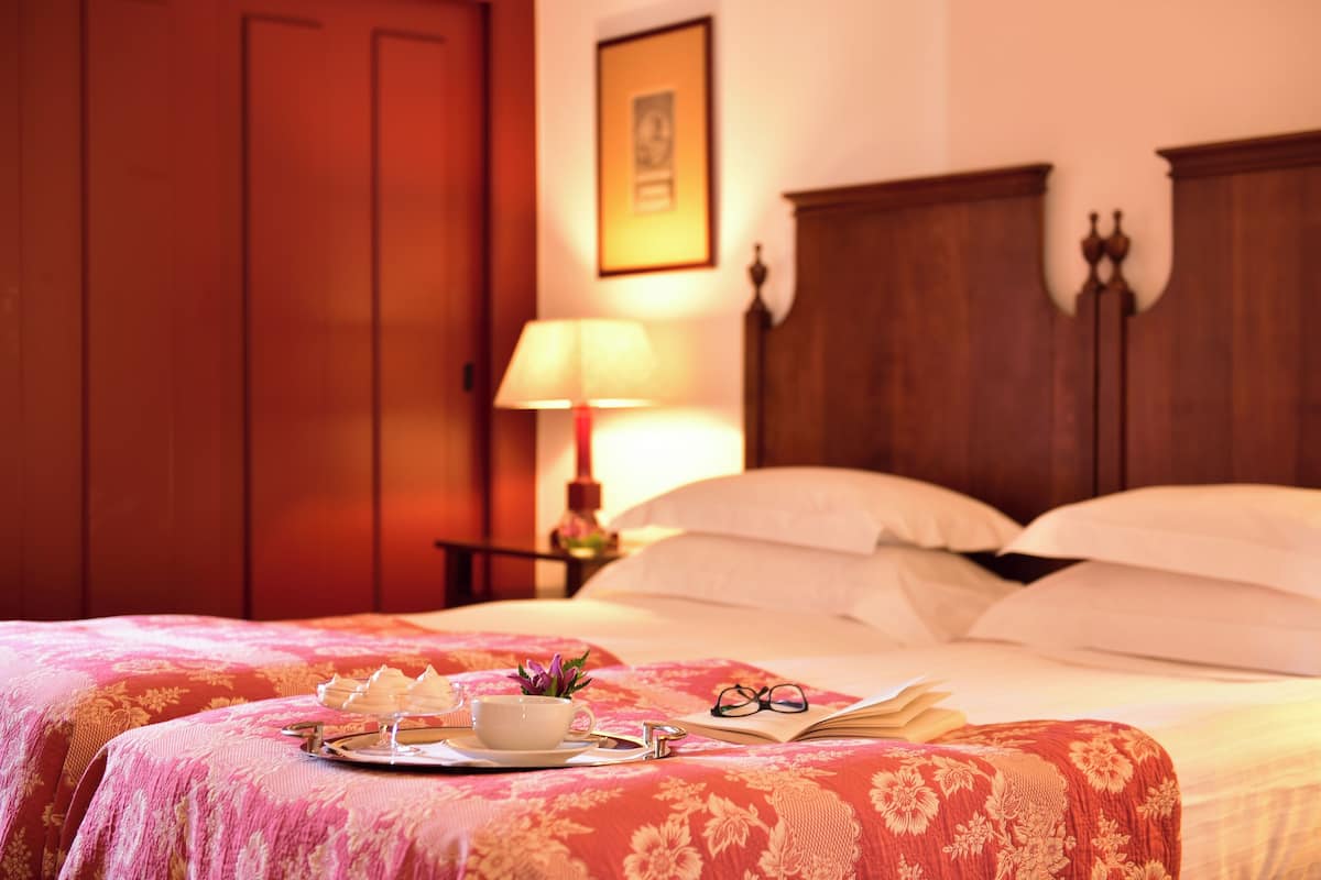 A neatly made bed with a floral bedspread, a tray with a cup and pastries, a book, and a pair of glasses on the bed.
