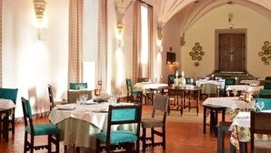 A dining area with tables set for service, chairs upholstered in teal, and a high arched ceiling with stone detailing.