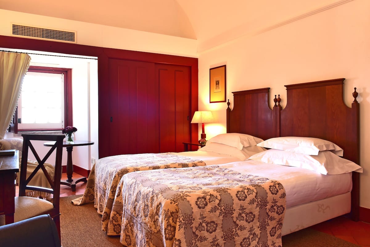 Quarto de hotel com duas camas, uma escrivaninha, uma cadeira, janela com cortinas e uma luminária fixada na parede.