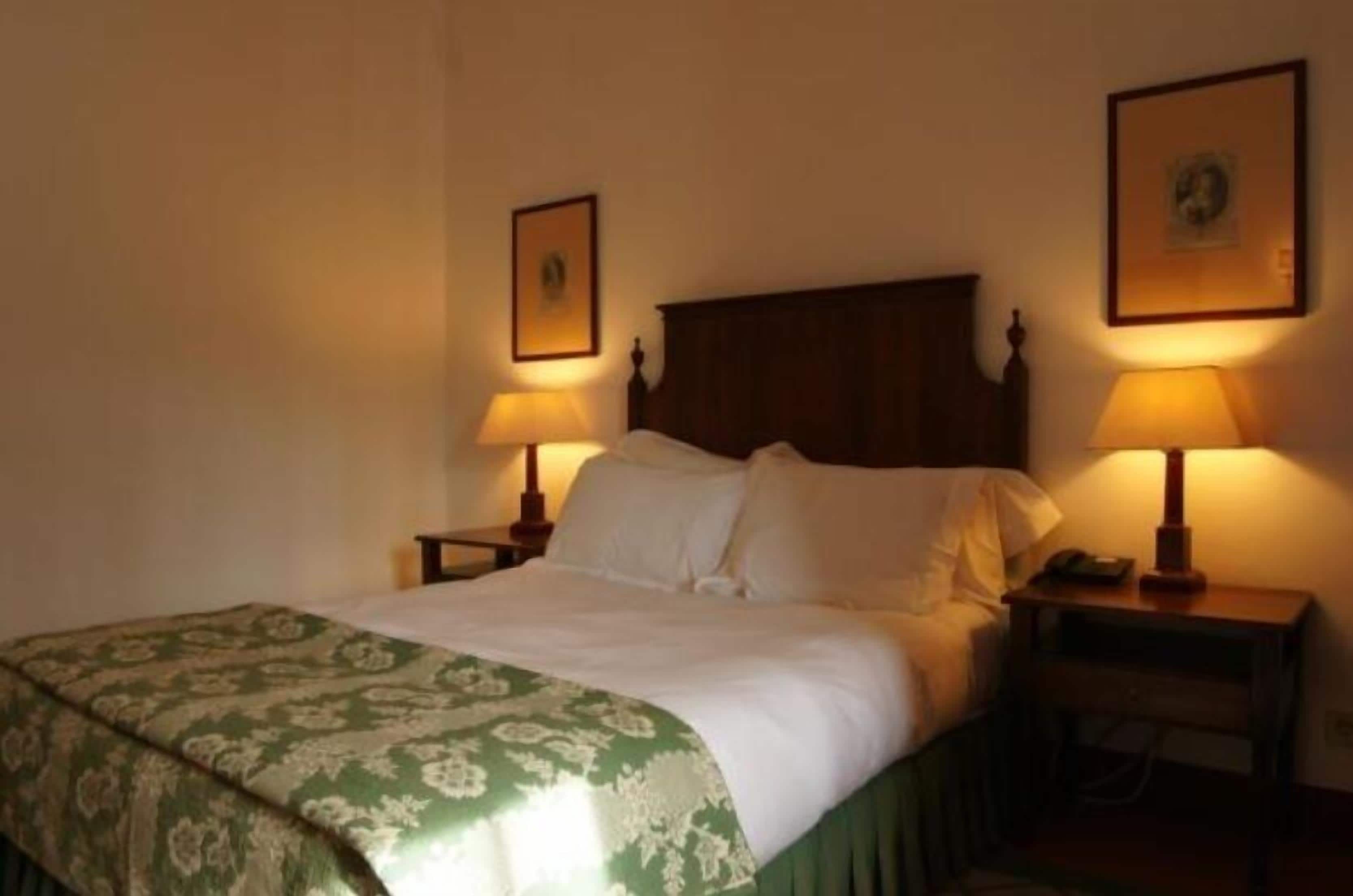 Quarto de hotel padrão com uma cama, duas mesas de cabeceira com abajures, um edredom verde estampado e duas imagens emolduradas nas paredes.