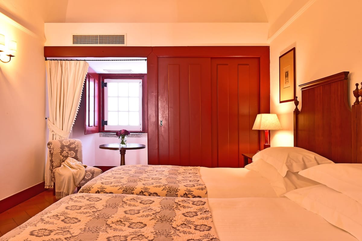 A hotel room with a large bed, a wooden wardrobe, a small table with a lamp, a chair, and a window with curtains.