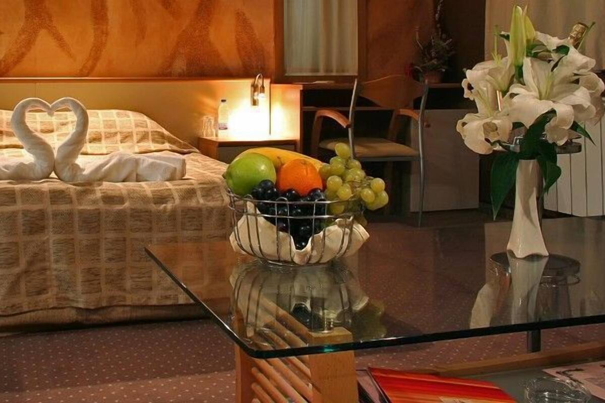 A hotel room with a bed, a glass coffee table with fruit and flowers, and a nightstand with a lamp.