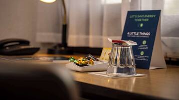 Ein Hotelzimmer mit Schreibtisch, Lampe, einem Glas mit rotem Deckel, einem Teller mit Essen und einem Schild zur Energiesparung.