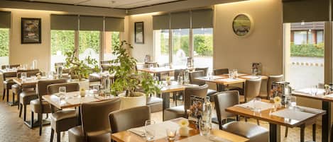 A restaurant with hexagonal tiled flooring, multiple tables with chairs, potted plants, and large windows offering a view of greenery outside.