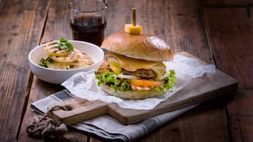 A burger on a wooden board with a napkin underneath, a glass of soda, and a bowl of fries on a wooden table.