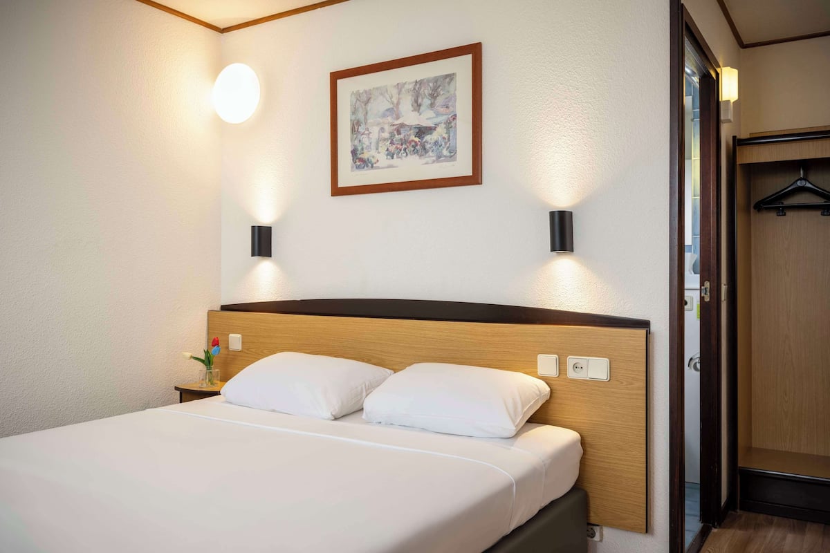 Hotellihuone, jossa on sänky, yöpöydällä olevat lamput, seinällä oleva taulu ja puinen vaatekaappi.