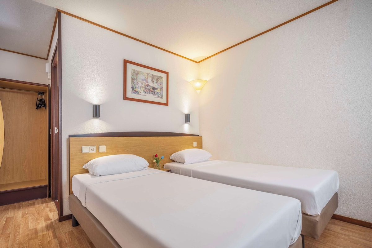 Hotellihuone, jossa on kaksi erillistä sänkyä, puinen pääty, seinällä kehystetty taulu ja puulattia.