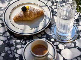 Um croissant em um prato com um framboeso e uma porção de geleia, um copo de água e uma xícara de café sobre uma mesa estampada.