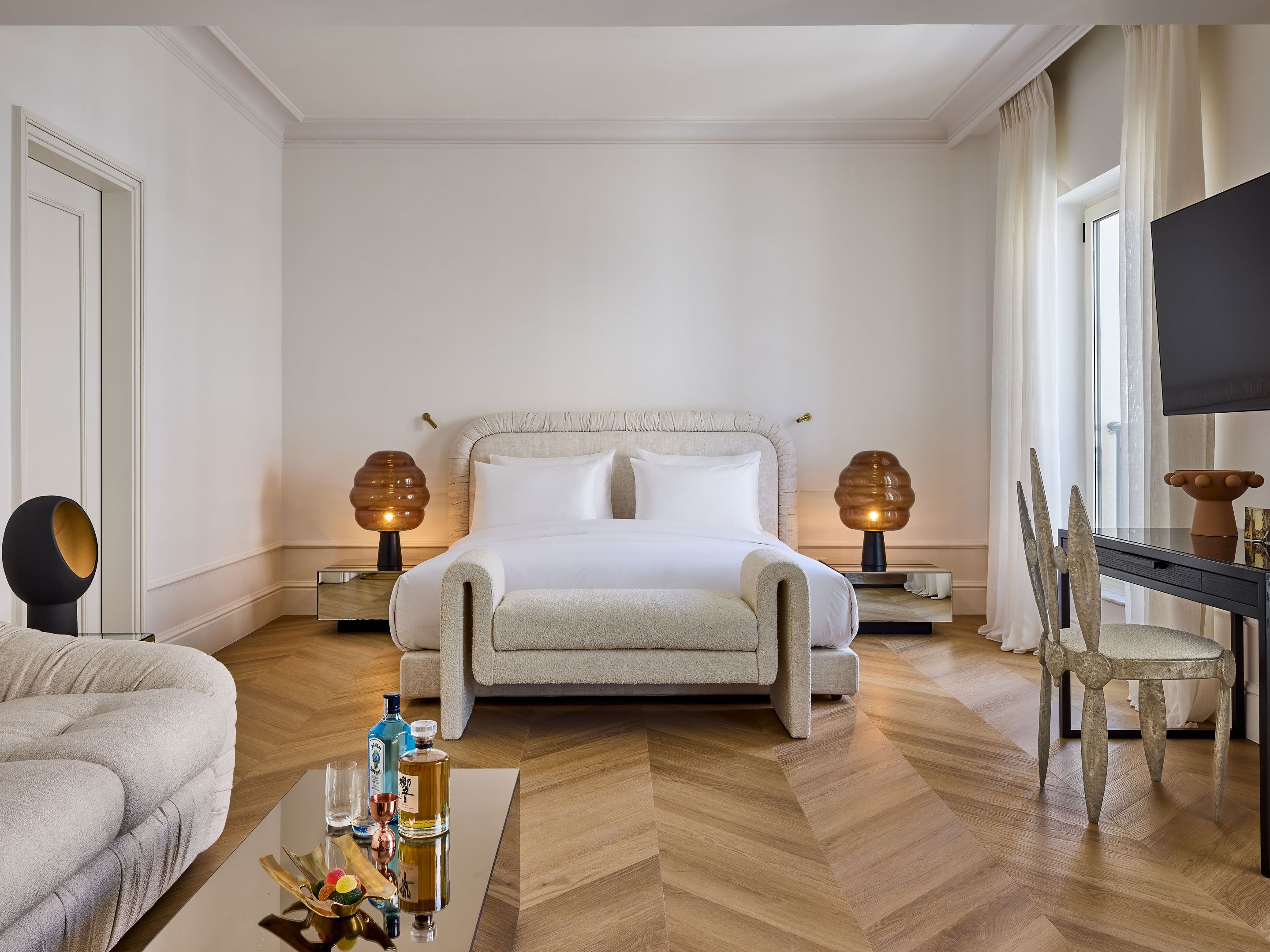Habitación de hotel con una cama grande, dos mesitas de noche con lámparas, un sofá, una mesa de comedor con sillas y un televisor.