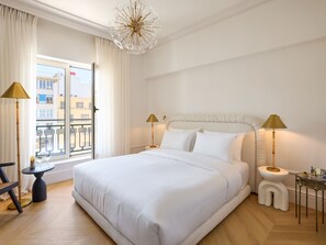 A hotel room with a large bed, two bedside lamps, a window with curtains, and a view of buildings outside. - ATHENA Pallas (Athens)