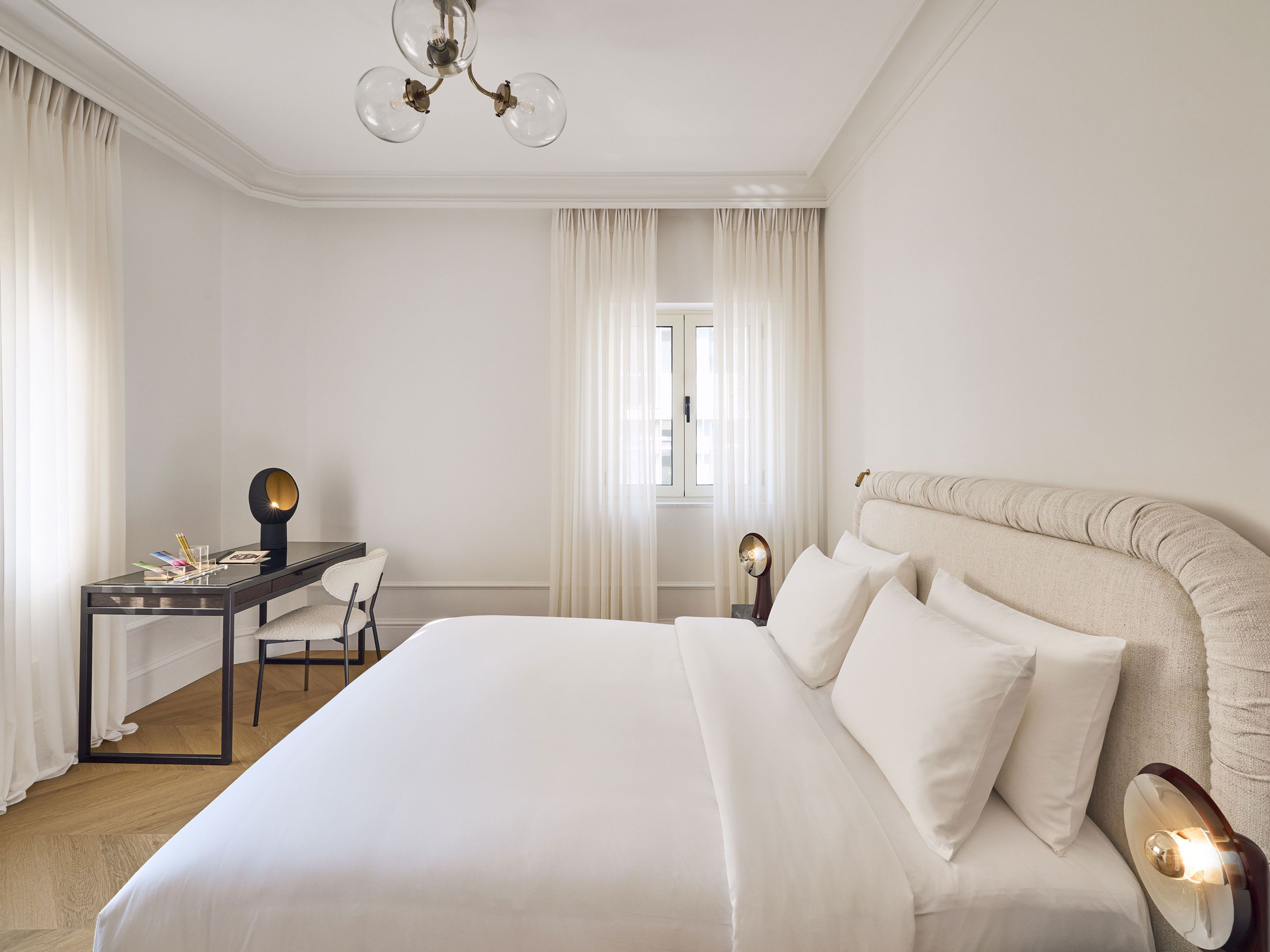 Een hotelkamer met een groot bed, een bureau en een stoel. Er is een raam met gordijnen, een kroonluchter en een bedlampje.