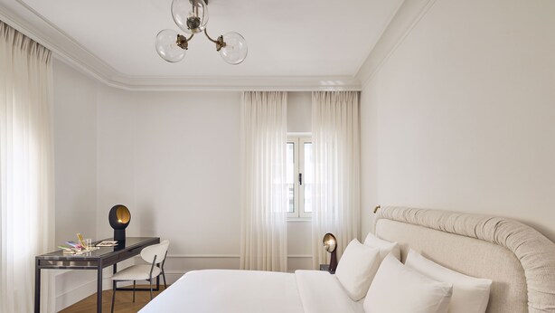 A hotel room with a large bed, a desk, and a chair. There is a window with curtains, a chandelier, and a bedside lamp. - ATHENA Pallas (Athens)