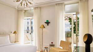 A hotel room with a large bed, a sitting area with a chair and table, a floor lamp, a mirror, and a view of the city through the window.