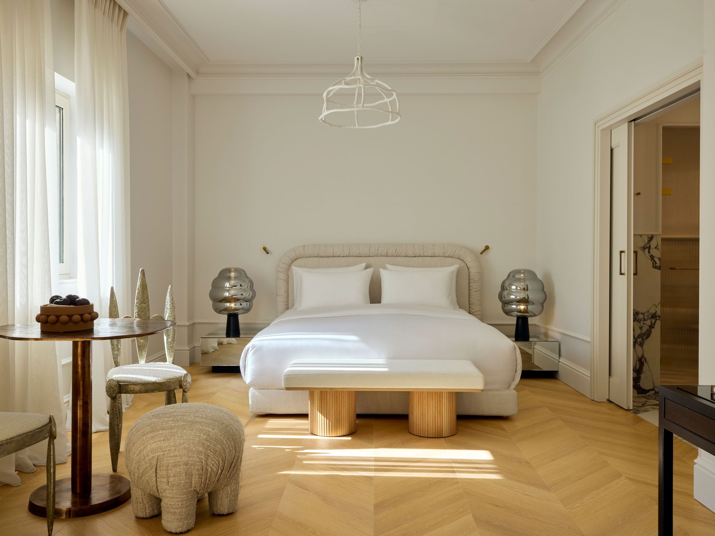 A bedroom with a large bed, bedside lamps, a round table with a bowl of fruit, a small stool, and a bench.