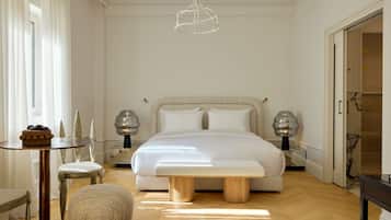 A bedroom with a large bed, bedside lamps, a round table with a bowl of fruit, a small stool, and a bench.