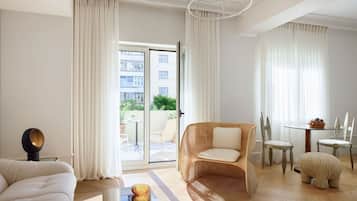 A modern hotel room with a sofa, a wicker chair, a small table with a bowl of fruit, and a view of the outside balcony.