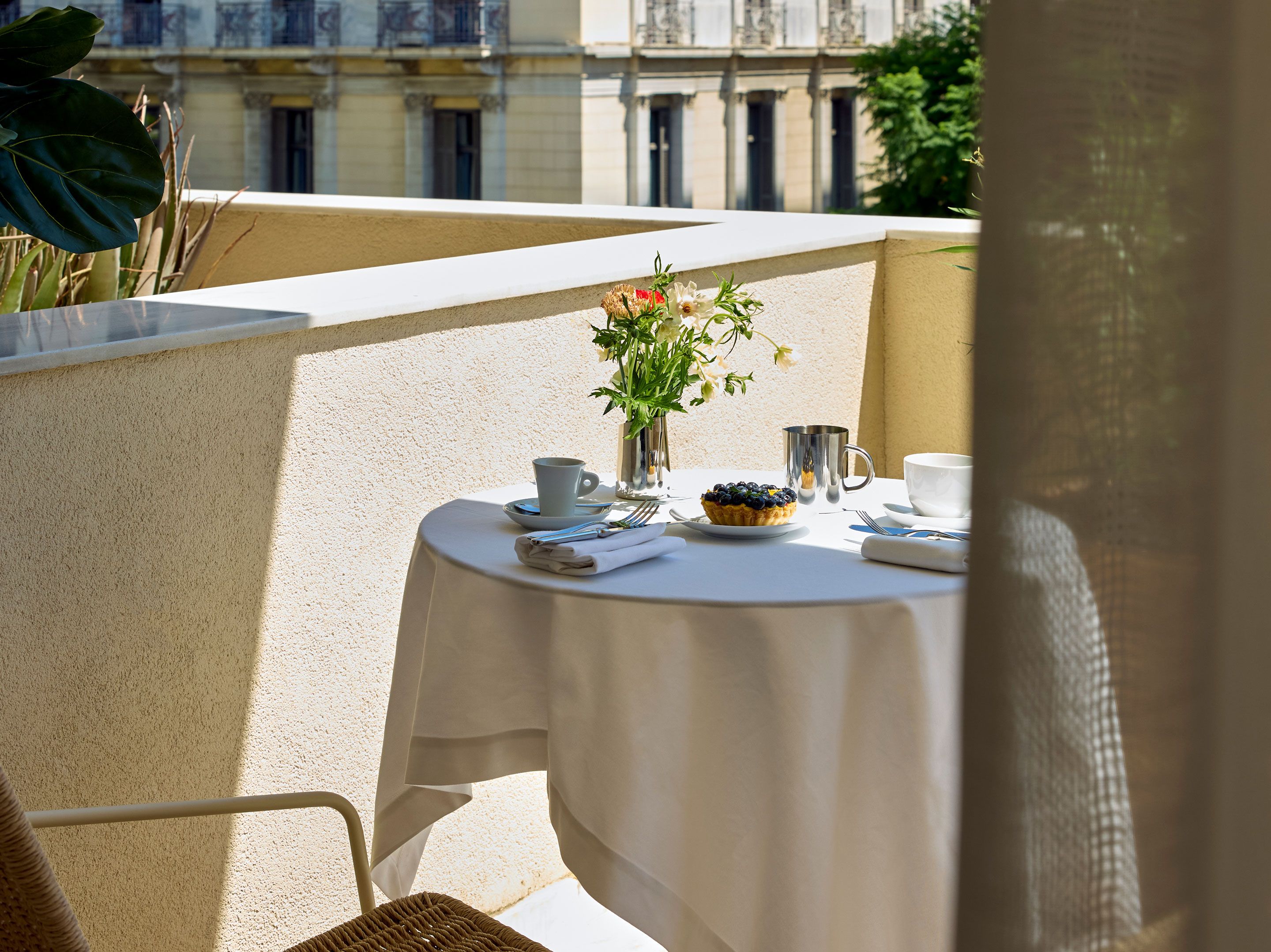 Un tavolo da balcone con una tovaglia bianca, un piccolo vaso con fiori, un piatto con una pasticceria e una tazza.