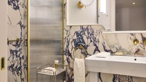 A bathroom with a marble wall, a glass shower enclosure, a sink with a gold faucet, and a mirror with a gold frame.