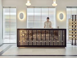 Un mostrador de recepción de diseño moderno, una persona de pie detrás y vistas al exterior a través de amplios ventanales.