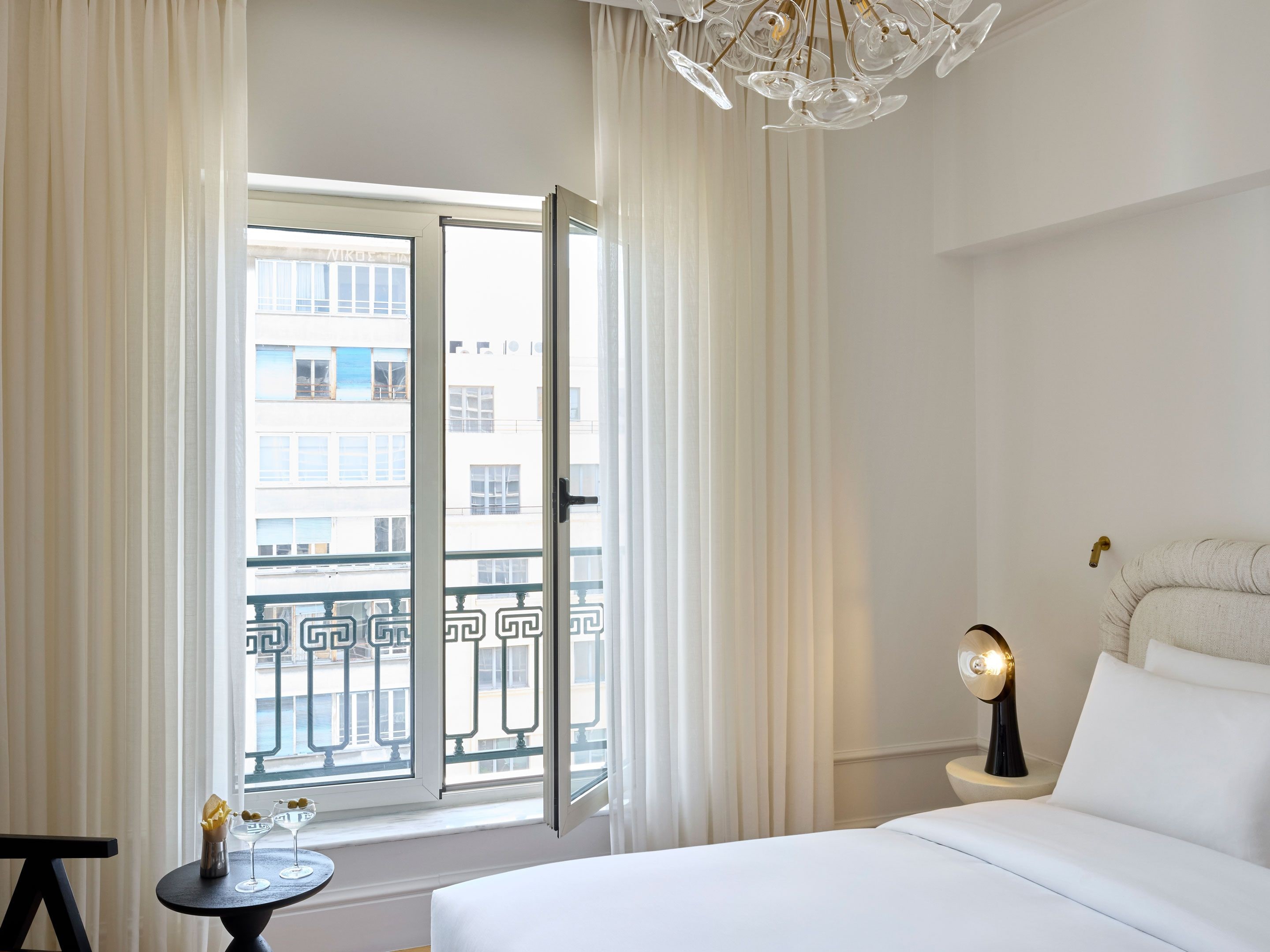 A hotel room with a bed, a nightstand, a table with drinks, and a view of the city through a balcony door.