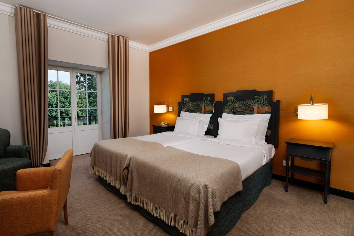 Habitación de hotel con una cama grande, dos mesitas de noche con lámparas, una silla y una ventana con cortinas.