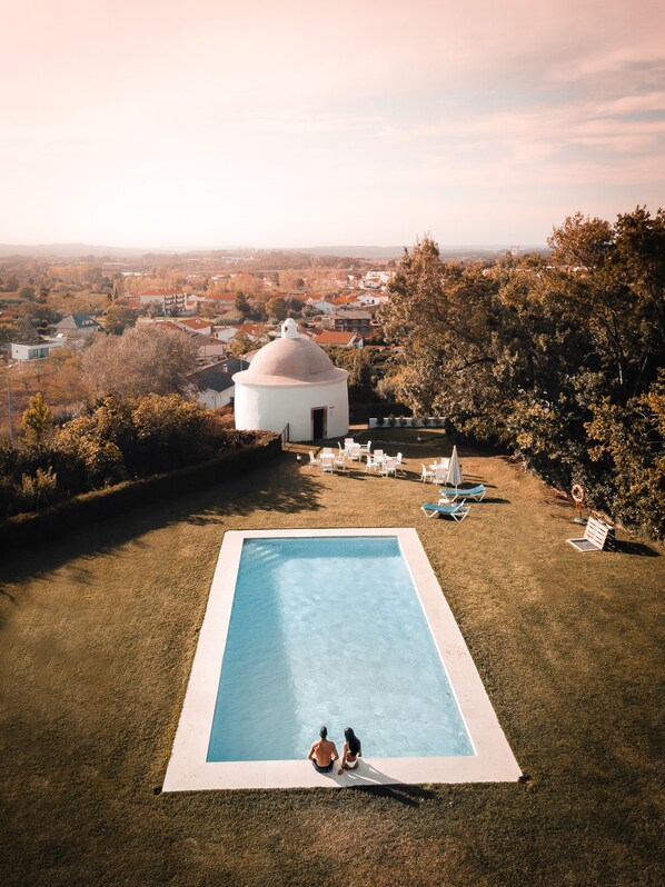 Una piscina con dos personas sentadas en el borde, rodeada de sillas de jardín y sombrillas, con un edificio blanco y árboles al fondo.
