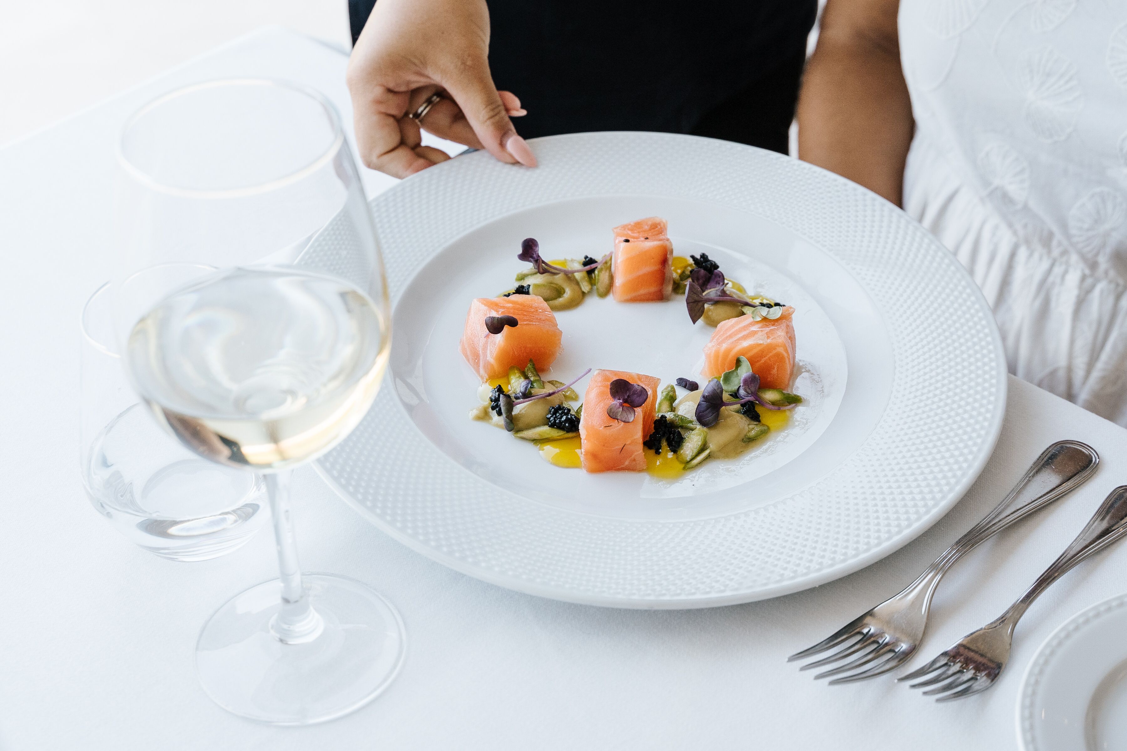 Un plato blanco con un plato de salmón, una copa de vino y cubiertos sobre un mantel blanco.