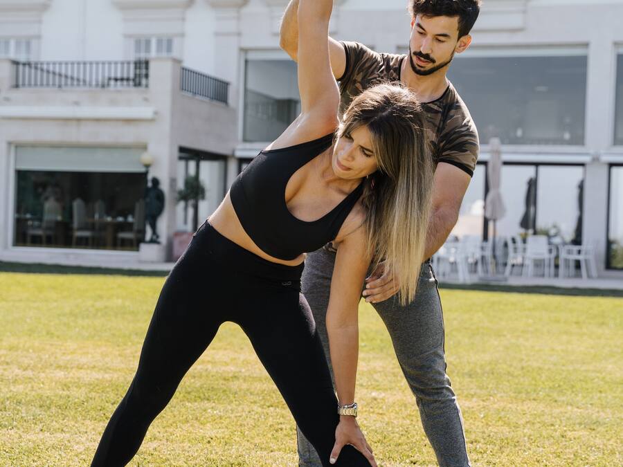 Dos personas practicando yoga sobre esterillas en un área verde al aire libre, con un edificio de fondo.