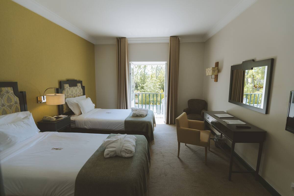 Habitación de hotel con dos camas, un escritorio, una silla, una lámpara y un balcón con cortinas.