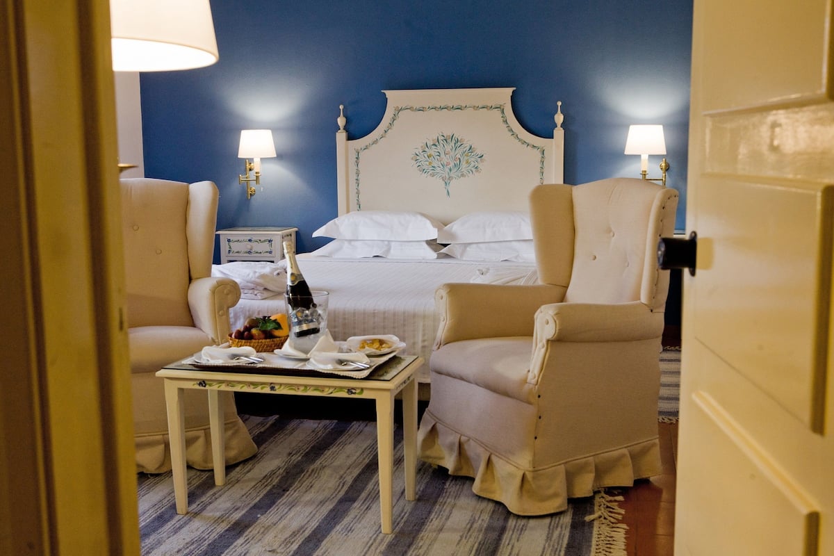 Une chambre d’hôtel avec un grand lit, deux lampes de chevet, une petite table avec un panier de fruits et deux fauteuils.
