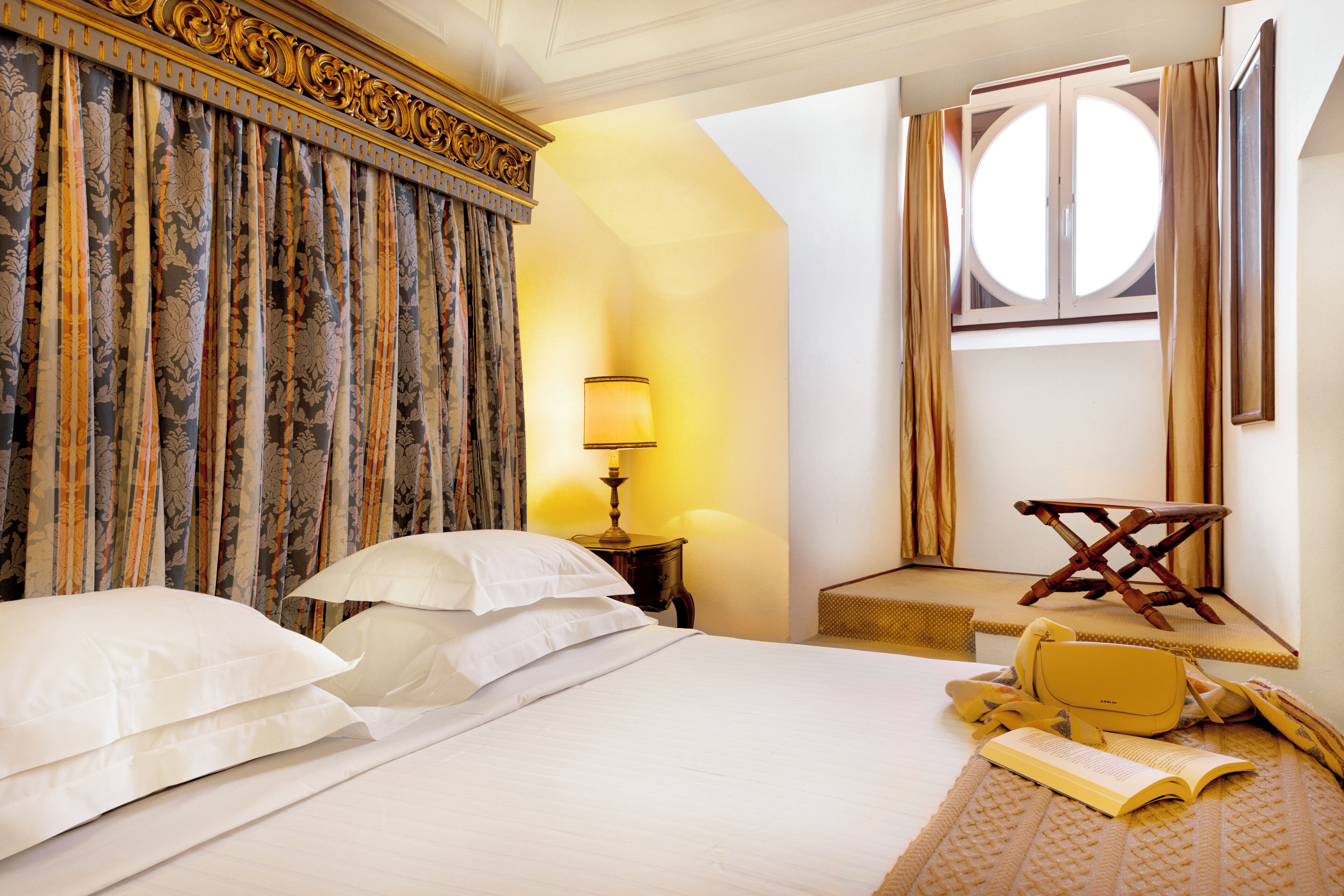 Una habitación de hotel con una cama grande, una mesita de noche con lámpara, una silla y una ventana con cortinas.