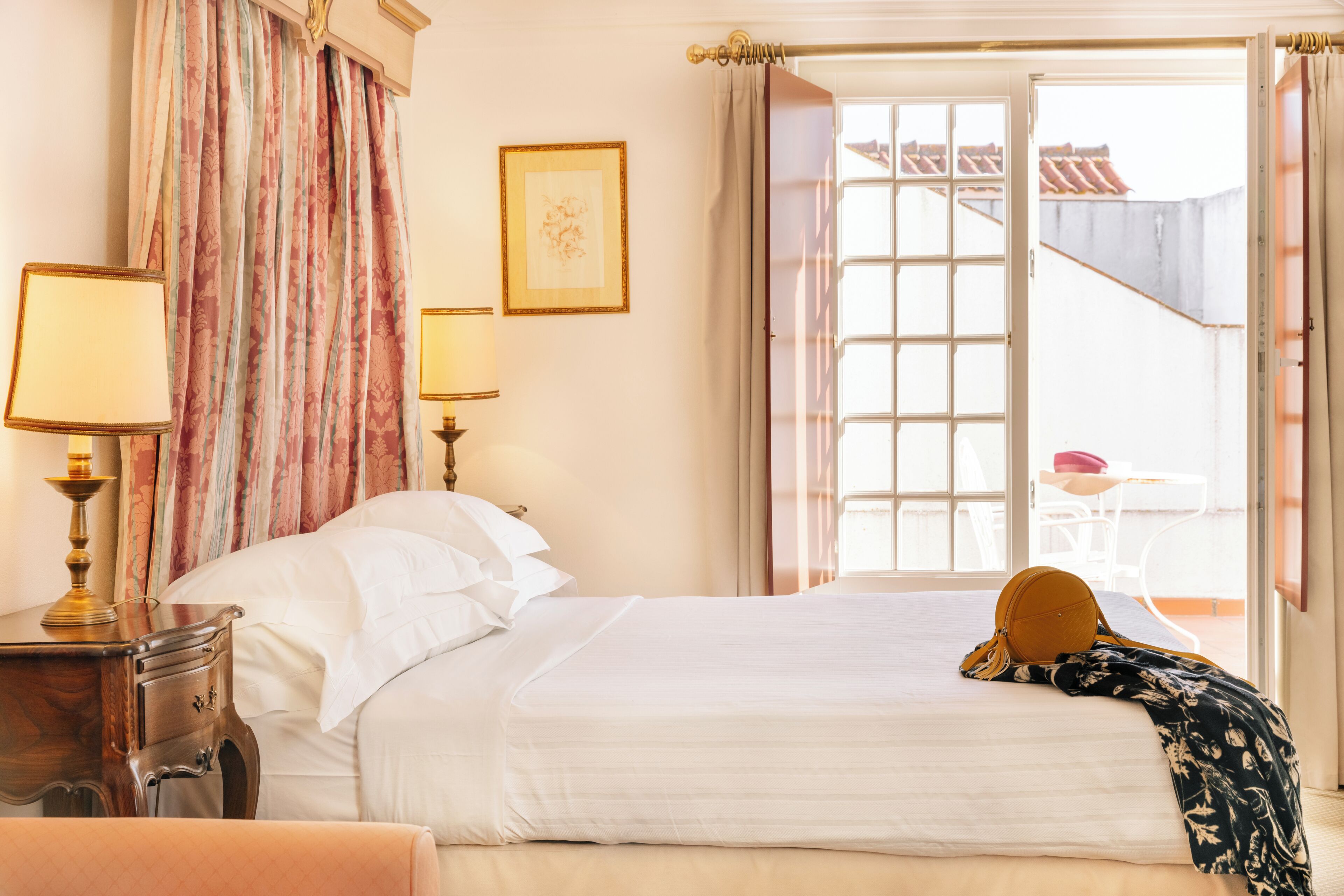 Una habitación de hotel con una cama, dos lámparas de noche, una ventana con cortinas y vistas al balcón exterior.