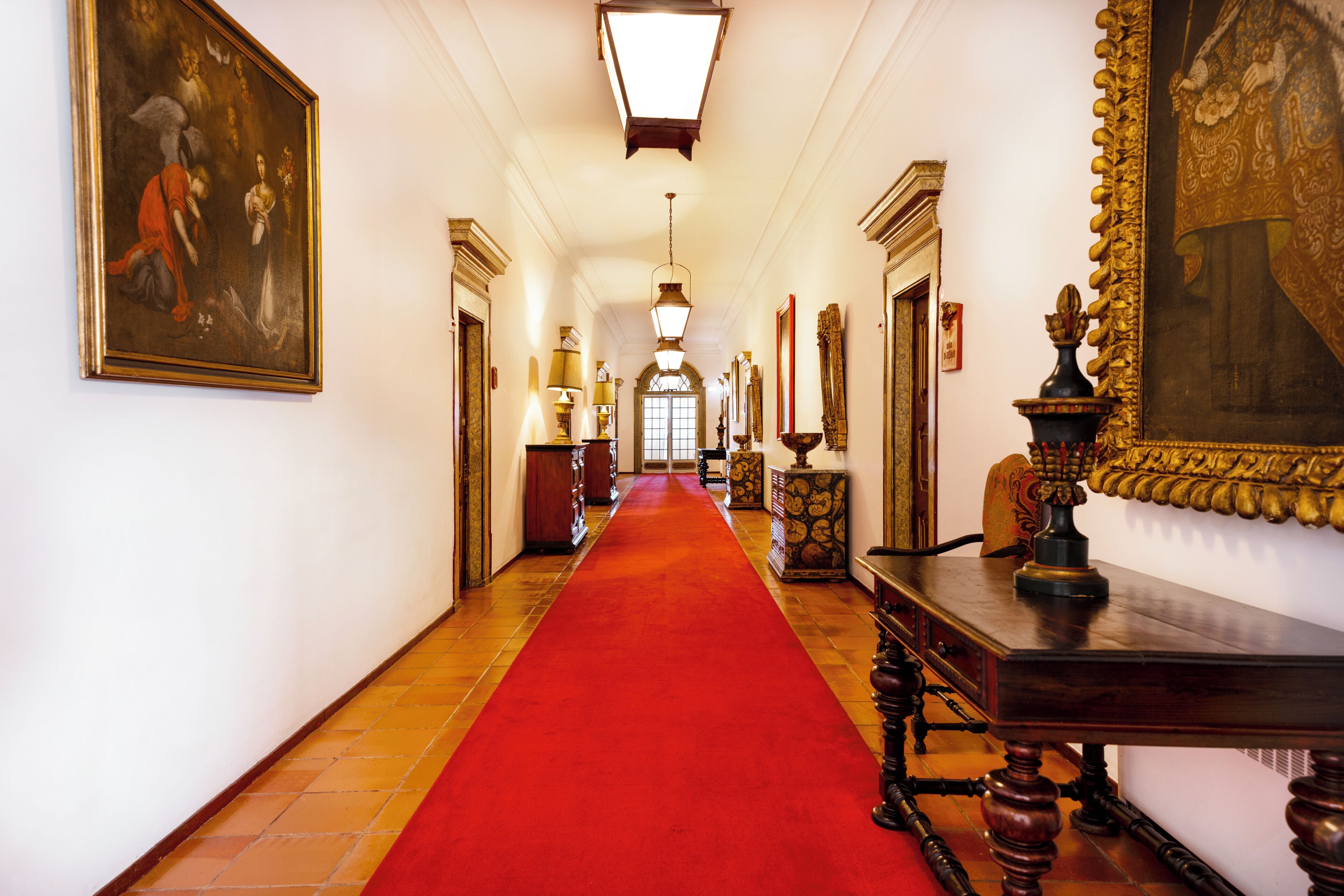 Un pasillo con alfombra roja, una araña de cristal y cuadros en las paredes.