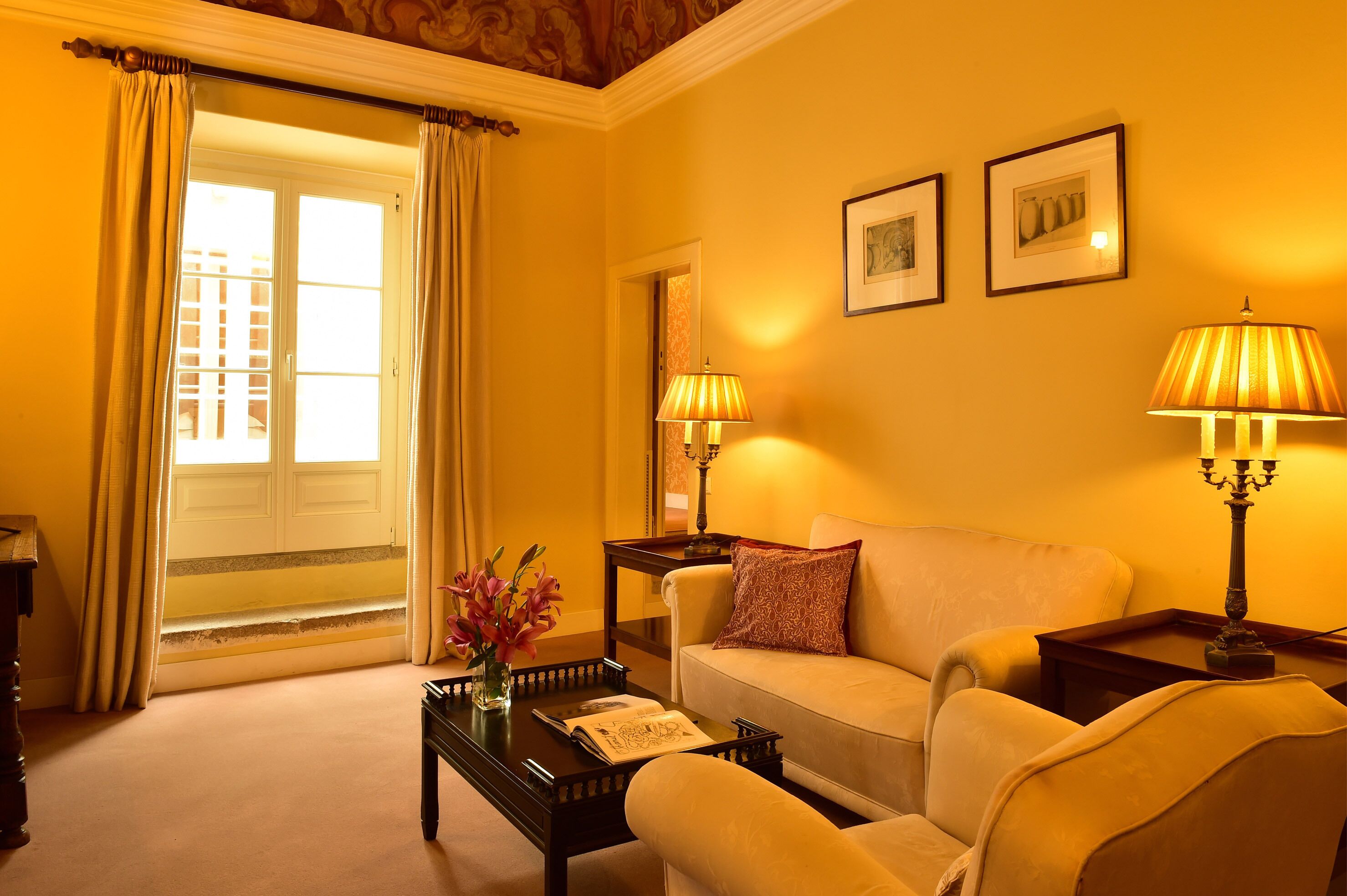 A well-lit living room with a sofa, two lamps, a coffee table with a vase of flowers, and a view of the outside through a door.