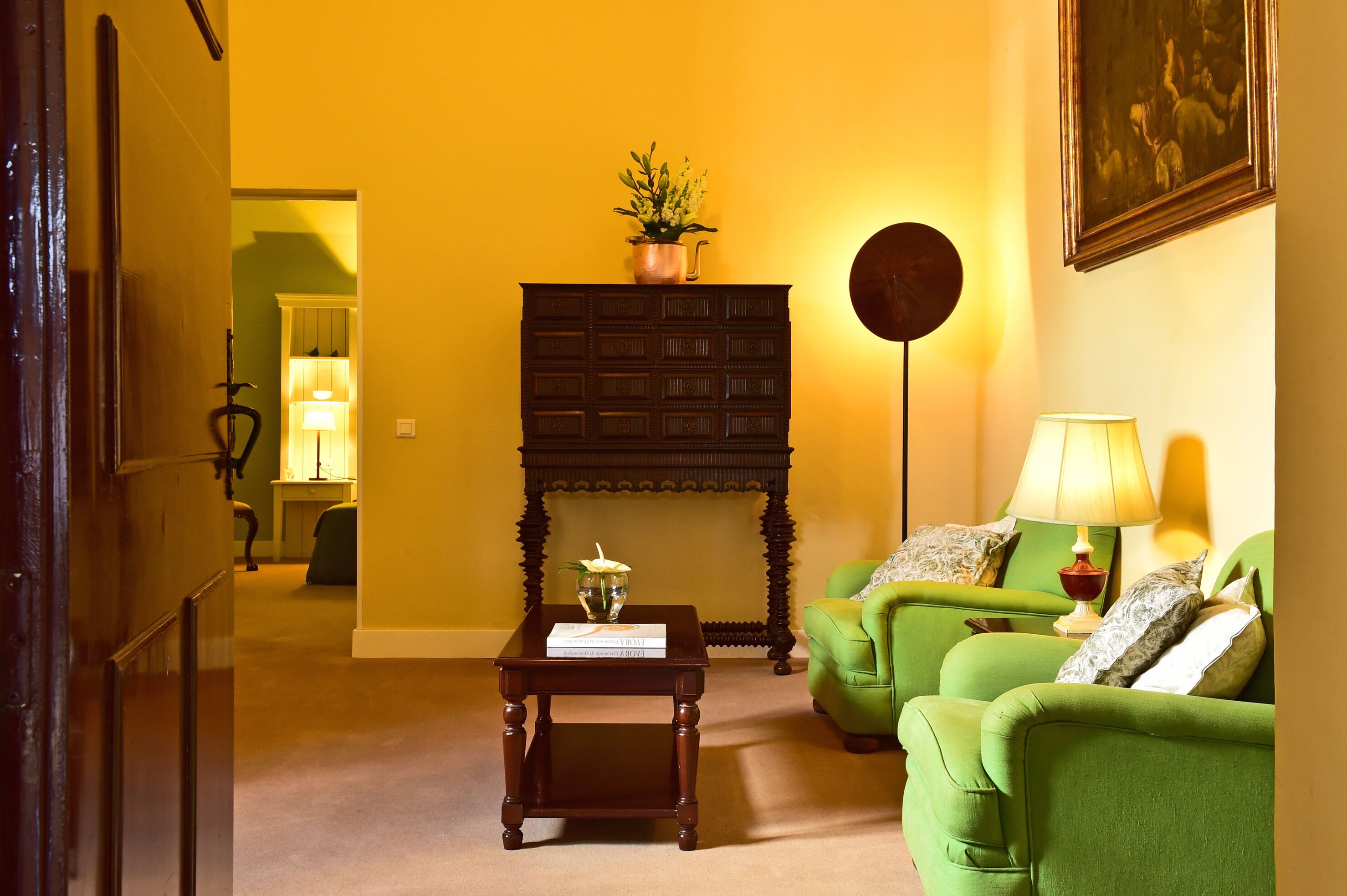 A room with a green sofa set, a dark wooden side table with a book, a small potted plant, and a wall-mounted round mirror.