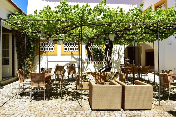 Un patio al aire libre con mobiliario de mimbre, una pérgola cubierta de hiedra y un suelo de adoquines.