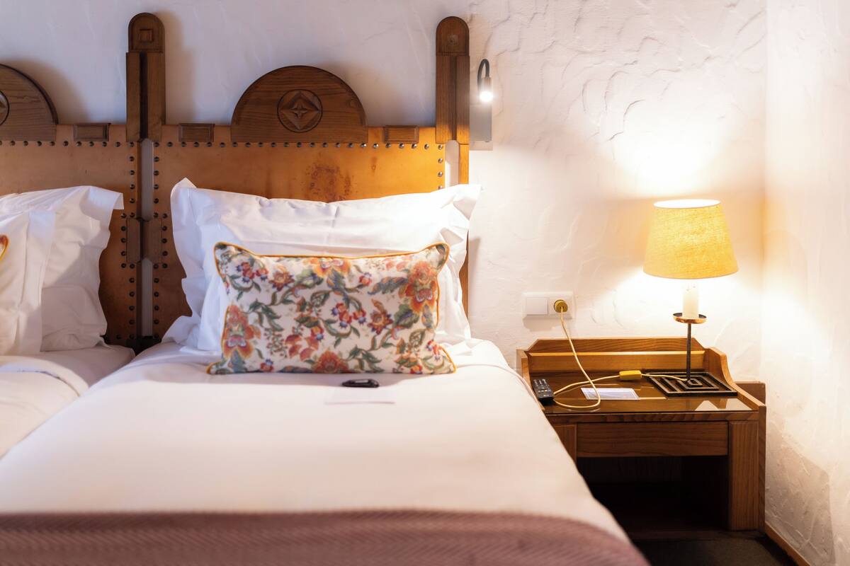 Een netjes opgemaakt bed met witte lakens en een bloemenkussen, een houten nachtkastje met een lamp, en een wandlamp.