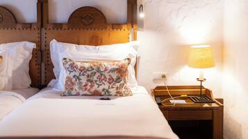 A neatly made bed with white linens and a floral pillow, a wooden nightstand with a lamp, and a wall-mounted light fixture.