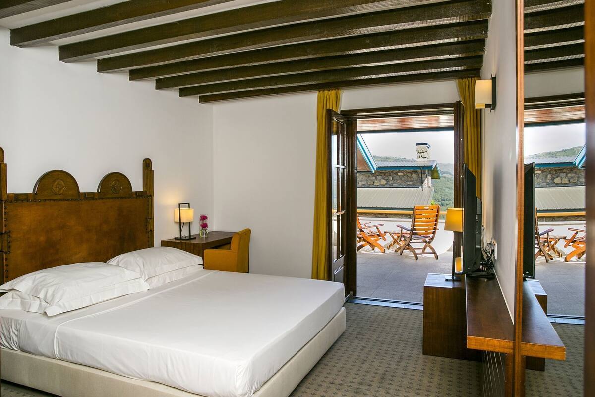 Een hotelkamer met een bed, een bureau, een stoel, een televisie en een balkon met zitgelegenheid buiten.