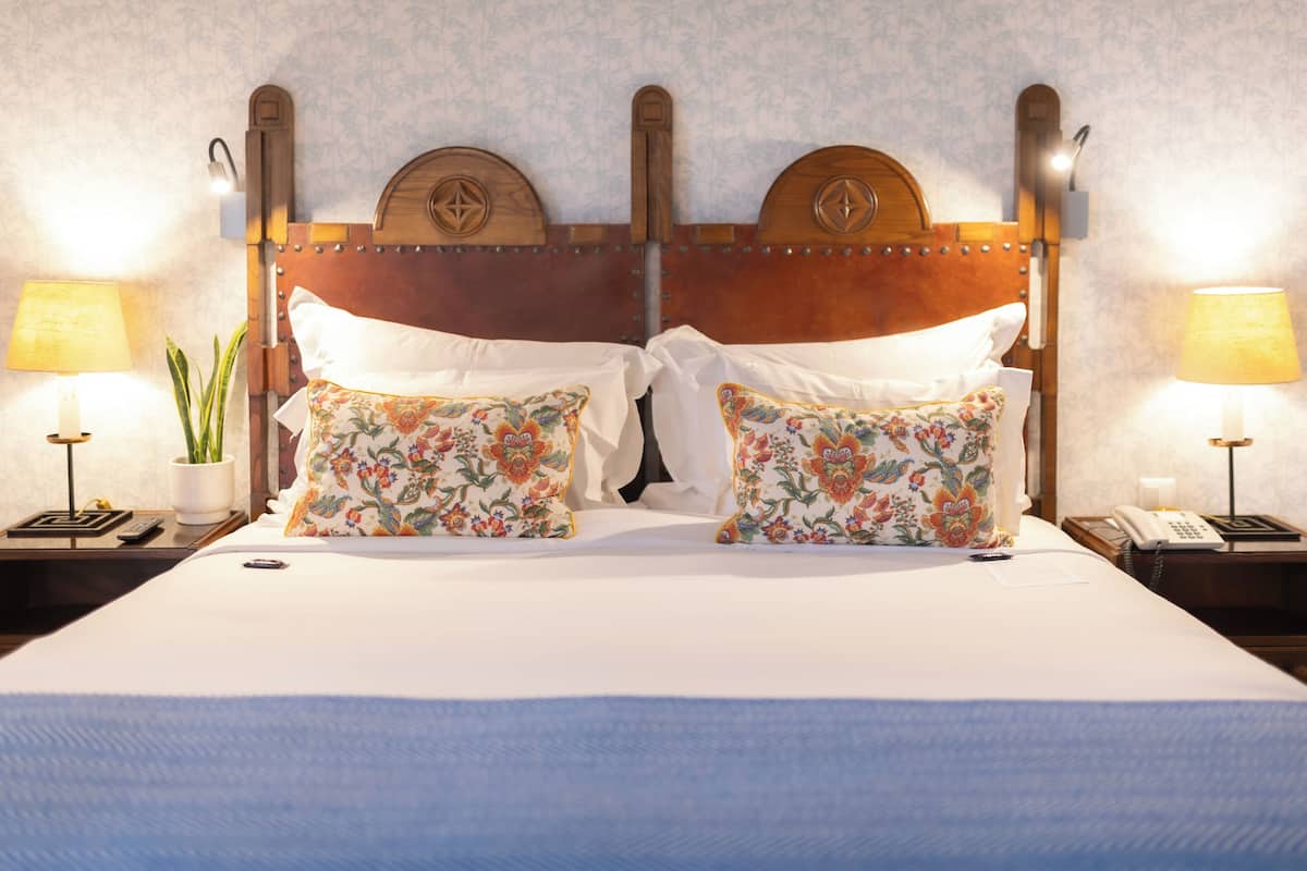 Een tweepersoonsbed met kussens met bloemenprint, een houten hoofdbord, bedlampjes, een nachtkastje met een telefoon en een plant, en een wand met een subtiel patroon.