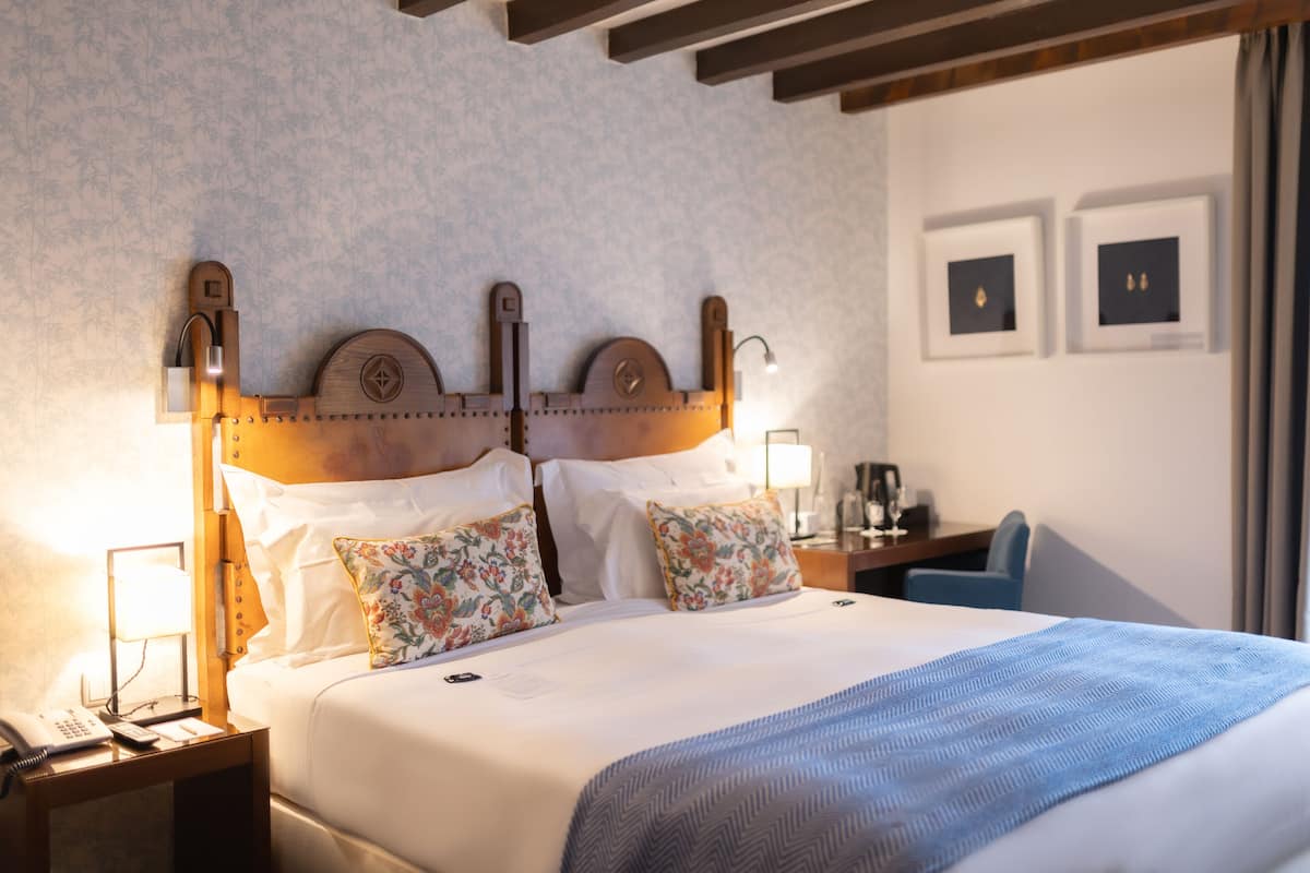 Een hotelkamer met een houten hoofdeinde, bloemenkussens, een blauw sprei, een nachtkastje met een lamp en ingelijste foto's aan de muur.